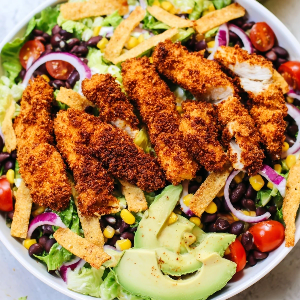 Crispy Chicken Taco Salad is shown in a white bowl, layered with romaine, black beans, corn, and golden chicken strips.