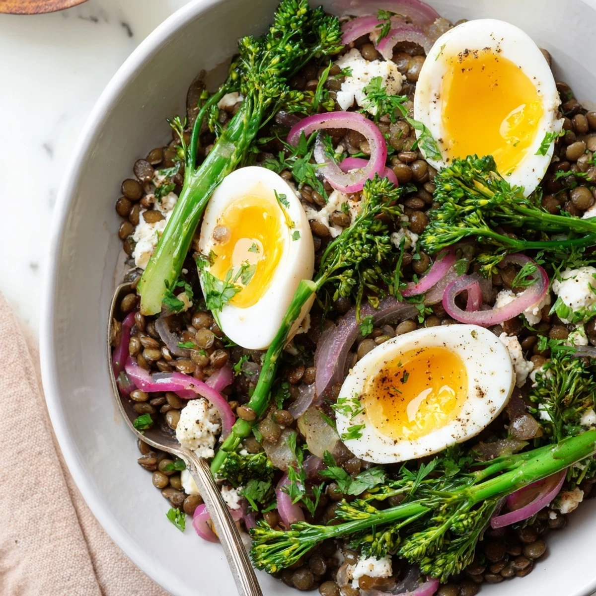 A close-up of French lentil salad with blanched broccolini and halved jammy soft-cooked eggs, drizzled with Dijon vinaigrette on a white plate.